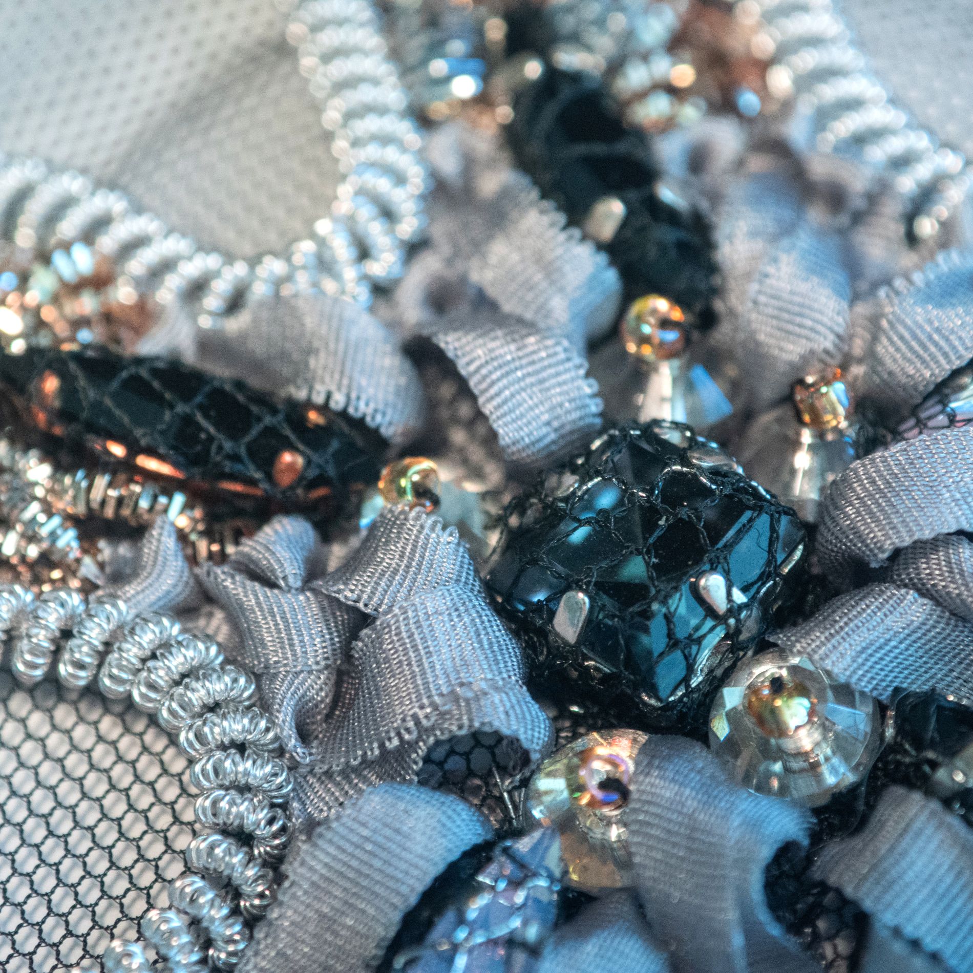 Couture Ribbon and Crystal Embroidery Detail Close-up of couture embroidery featuring folded grey ribbon, faceted crystals, metallic beads, and black beads wrapped in netting on a mesh base.