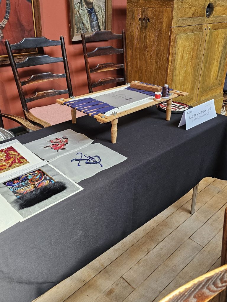 Table display by Ksenia Semirova showing a Luneville embroidery frame with work in progress, threads, and multiple couture-inspired embroidery samples at the Society of Designer Craftsmen group meeting, May 2025.
