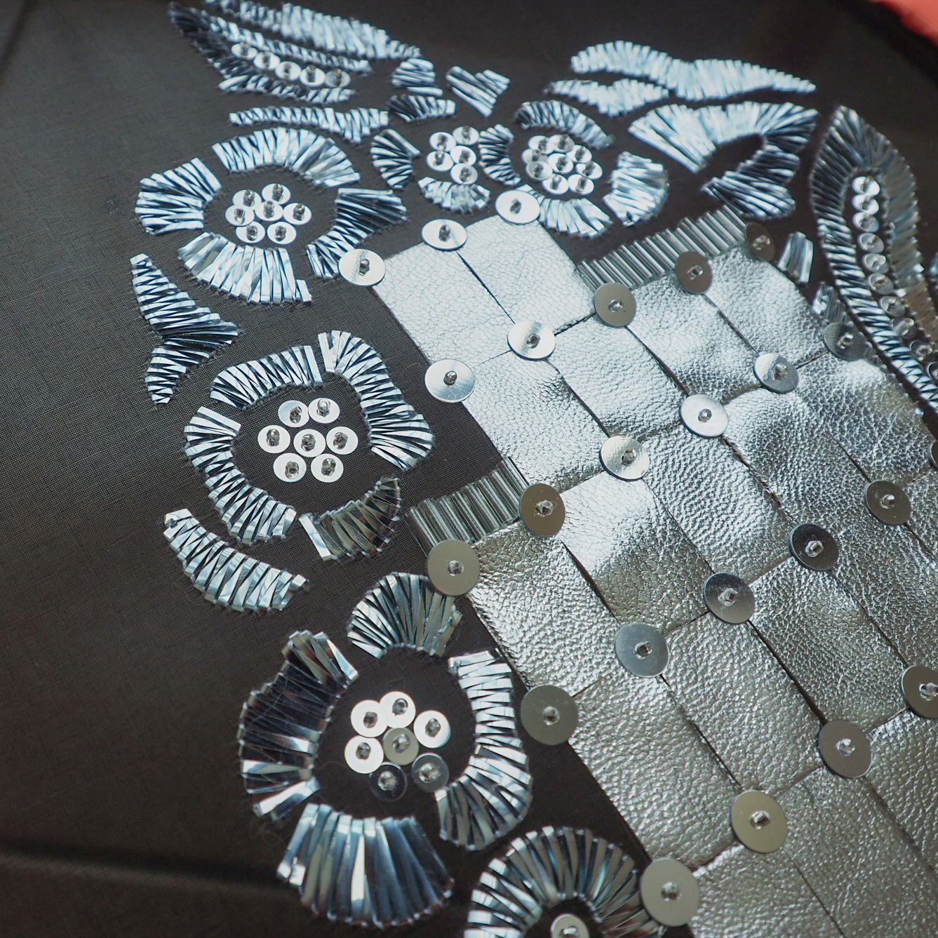 Schiaparelli Metallic Embroidery Study – Silver Paillettes & Foil Work Close-up of a couture embroidery sample inspired by Schiaparelli, featuring silver paillettes, cut metallic foil elements, and interlinked silver squares stitched on black fabric.