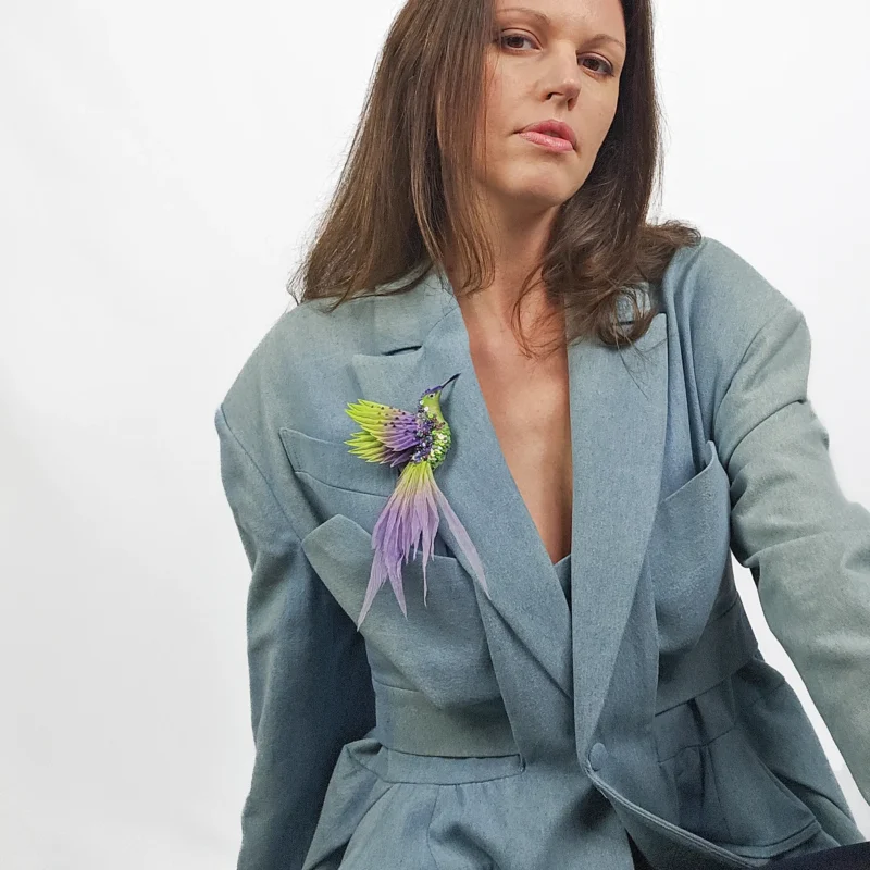 A woman wearing a light blue suit poses with the "Flying Hummingbird in Lime & Purple: Luxury Brooch" pinned to the lapel. The brooch showcases vibrant lime green, yellow, and purple details with intricate feathered accents, adding an elegant and artistic touch to the outfit. The background is plain white