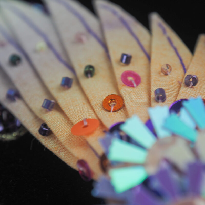 Close-up of the Hummingbird Spreading Wings luxury brooch, showcasing intricate embroidery with vibrant feathers, shimmering beads, and colorful accents against a black background