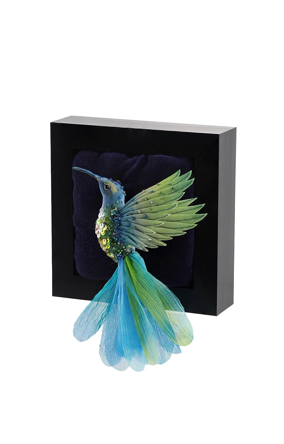 Handcrafted hummingbird brooch in aqua, green, and blue tones. The brooch is made from hand-dyed silk and cotton with intricate beadwork. It is displayed in a shadow box as wall art.