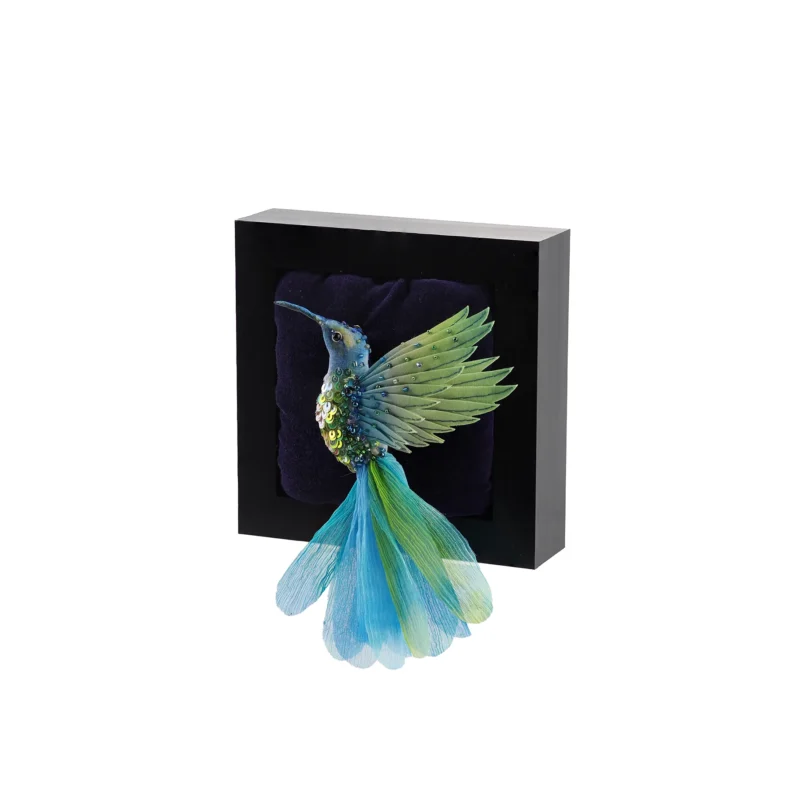 Handcrafted hummingbird brooch in aqua, green, and blue tones. The brooch is made from hand-dyed silk and cotton with intricate beadwork. It is displayed in a shadow box as wall art.