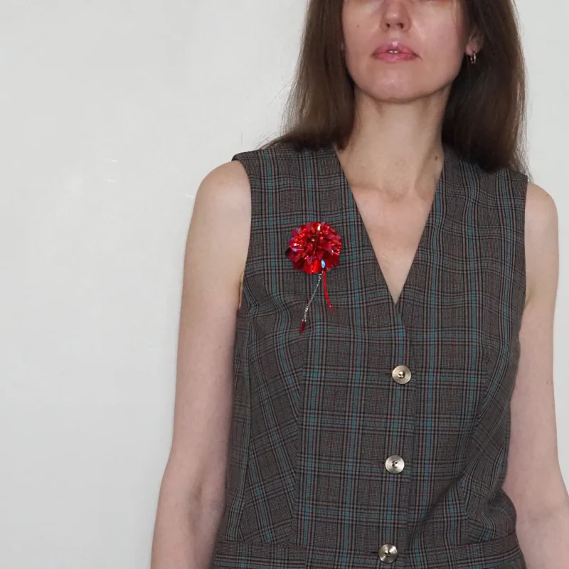 Model wearing a plaid sleeveless vest with a handcrafted red dahlia brooch pinned to the left side. The brooch features intricate beadwork with a blood drop bead and delicate chain accents, adding a striking, luxurious touch to the outfit. This bold accessory is designed to appeal to gothic style admirers and emphasizes the brooch's exclusivity and craftsmanship