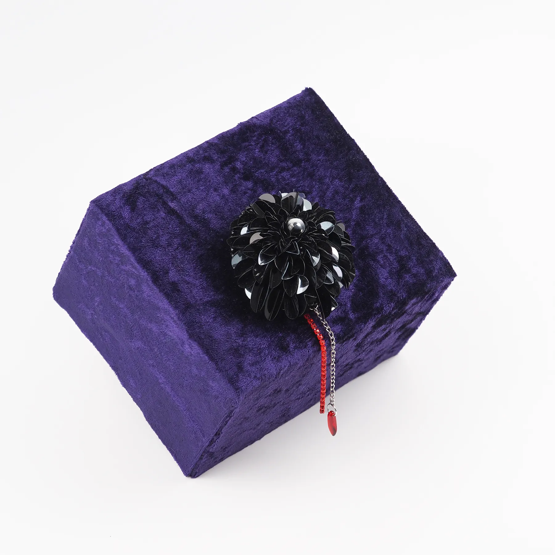 A handcrafted black dahlia brooch featuring layered black sequins, a central bead, and a dangling red rhinestone chain detail resembling a blood drop. The brooch is displayed on a deep purple velvet box, highlighting its Gothic-inspired design and dramatic elegance