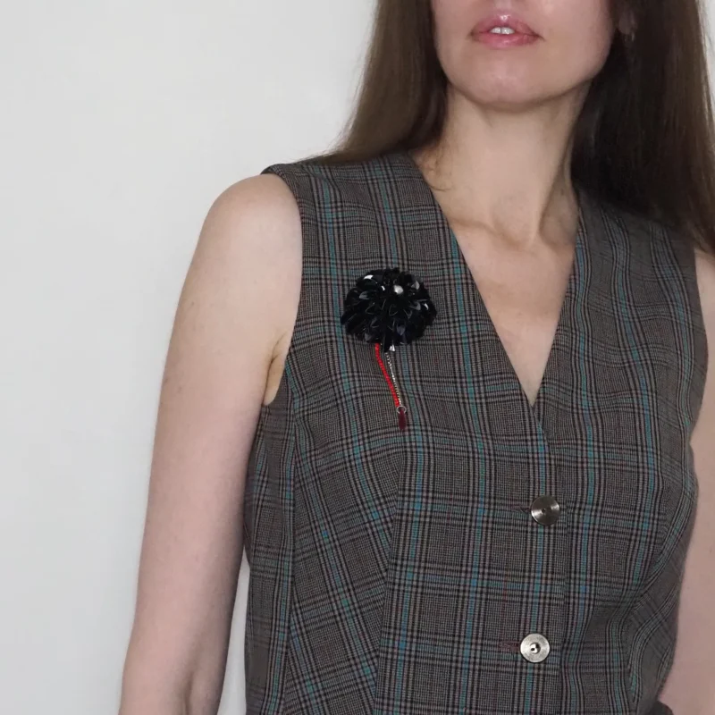 Model wearing a plaid sleeveless vest with a handcrafted black dahlia brooch pinned to the left side. The brooch showcases intricate beadwork with a blood drop bead and a delicate chain accent, creating a sophisticated, gothic-inspired accessory. This unique brooch adds a bold statement to any outfit and emphasizes exclusivity and high-quality craftsmanship