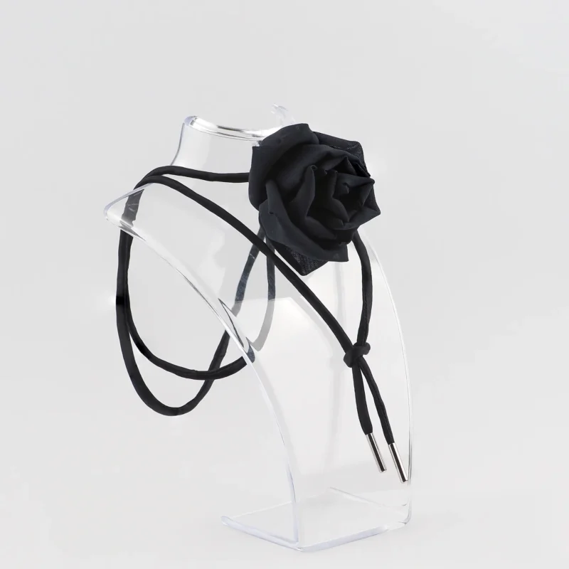 Black silk rose choker by Ksenia Semirova displayed on a clear acrylic stand. This handcrafted accessory features a black silk rose centerpiece, black satin lace, and nickel-free metal ends, highlighting elegant design and artisanal craftsmanship.