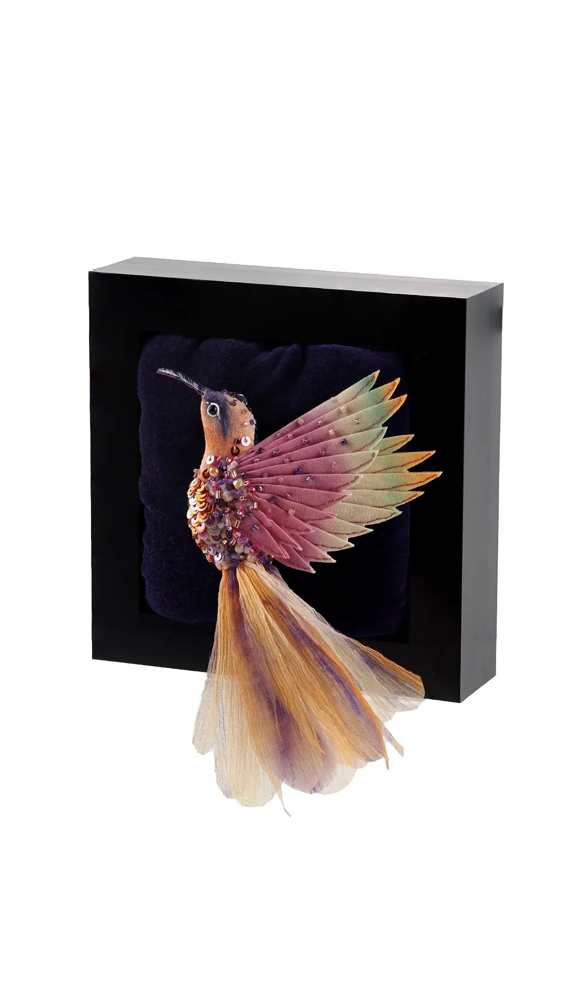 Flying Hummingbird Brooch in pastel pink, purple, and orange, set in a black display frame. The brooch showcases delicate silk wings, shimmering beads, and a graceful silk tail.