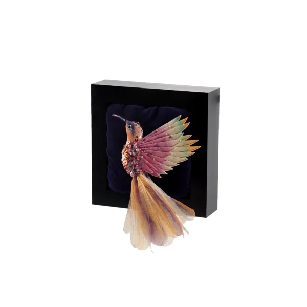 Flying Hummingbird Brooch in pastel pink, purple, and orange, set in a black display frame. The brooch showcases delicate silk wings, shimmering beads, and a graceful silk tail.