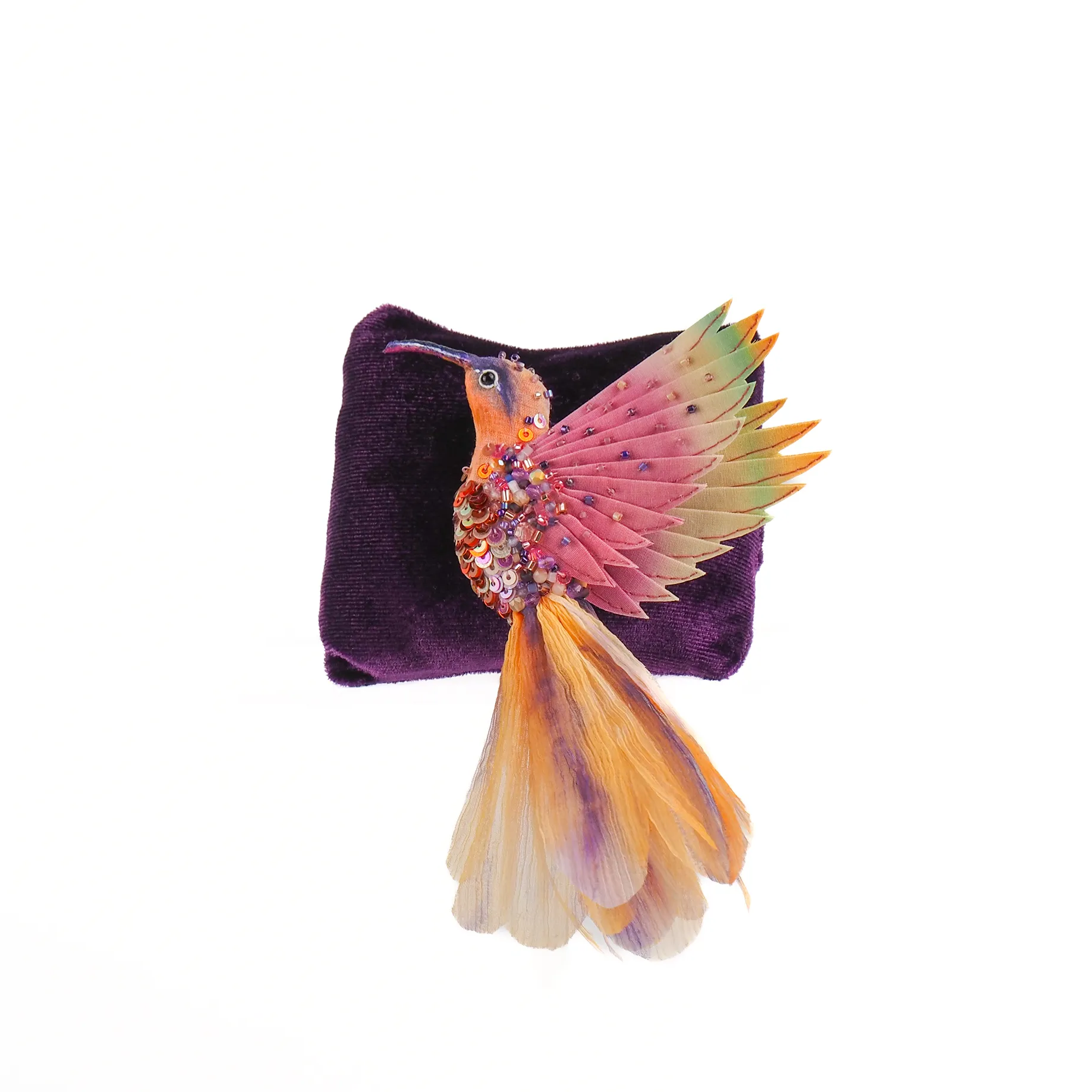 Flying Hummingbird Brooch in pastel pink, purple, and orange. The brooch features layered silk wings, intricate beadwork, and a flowing tail, displayed on a purple fabric backing.