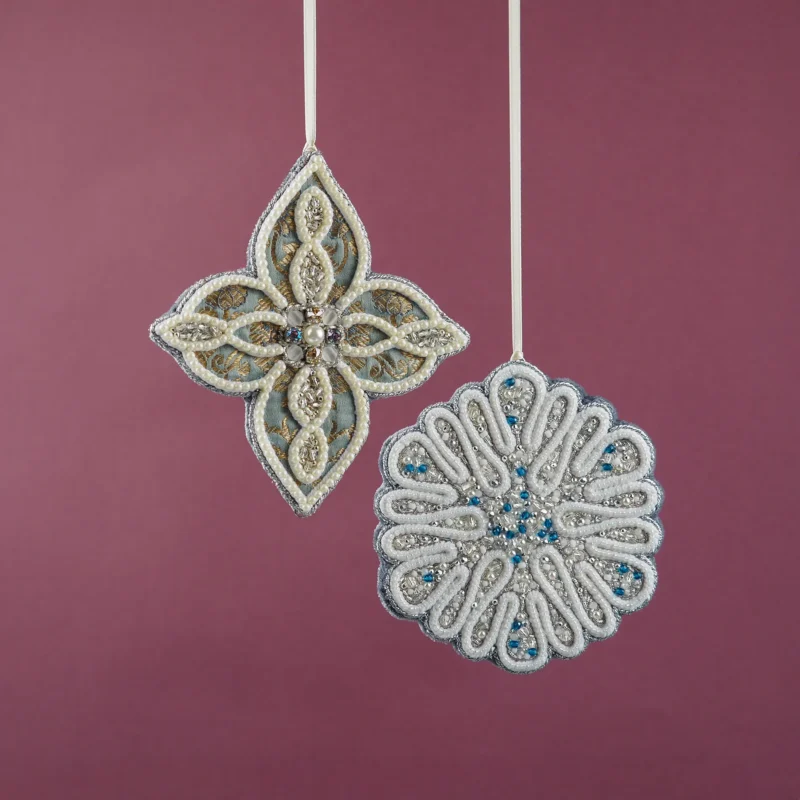 Elegant hand-embroidered ornaments with intricate beadwork and pearls, featuring a floral and snowflake-inspired design on fabric with gold and silver accents, displayed against a mauve background