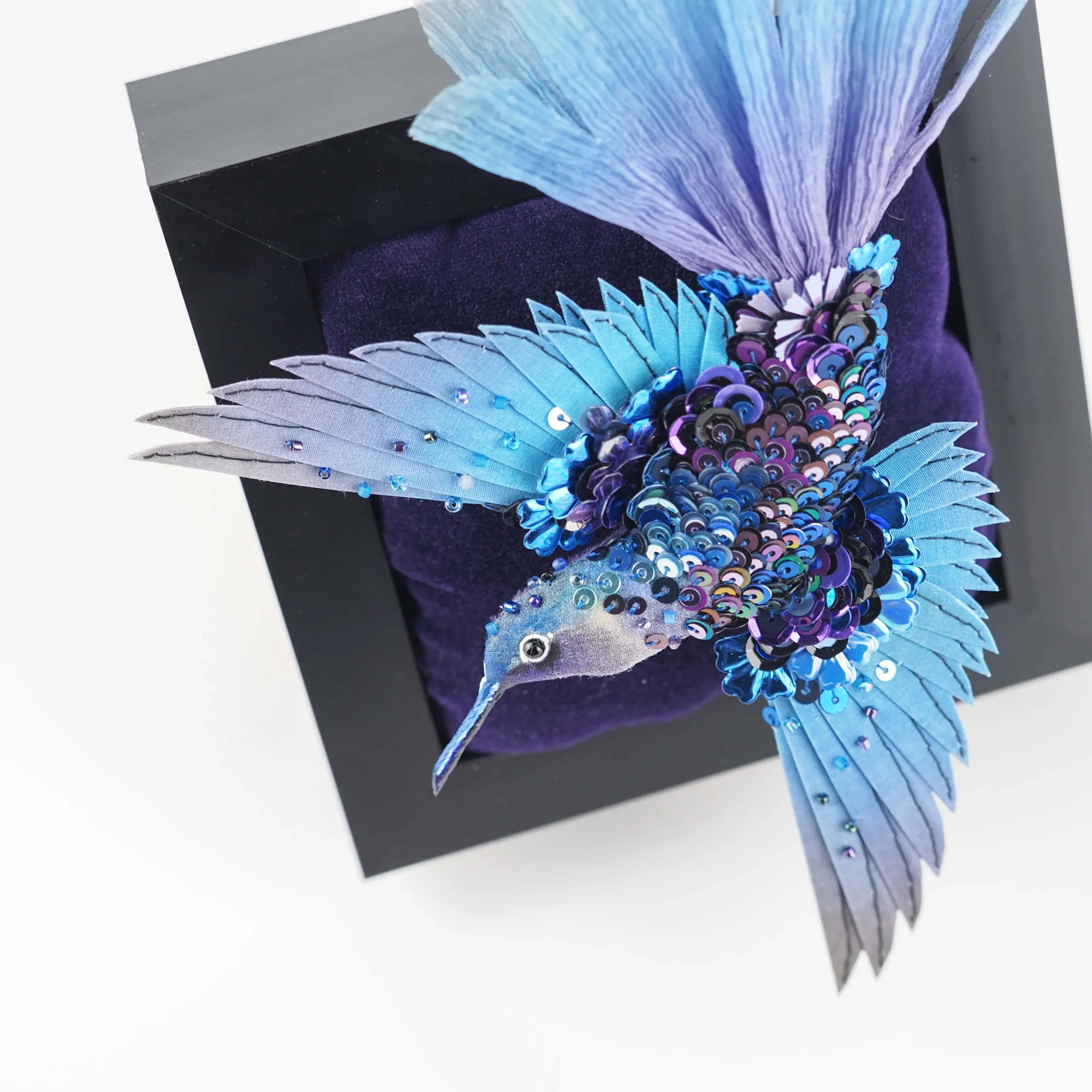 A luxury handcrafted hummingbird brooch with shimmering blue sequins and feathers, displayed on a deep velvet background in a black frame