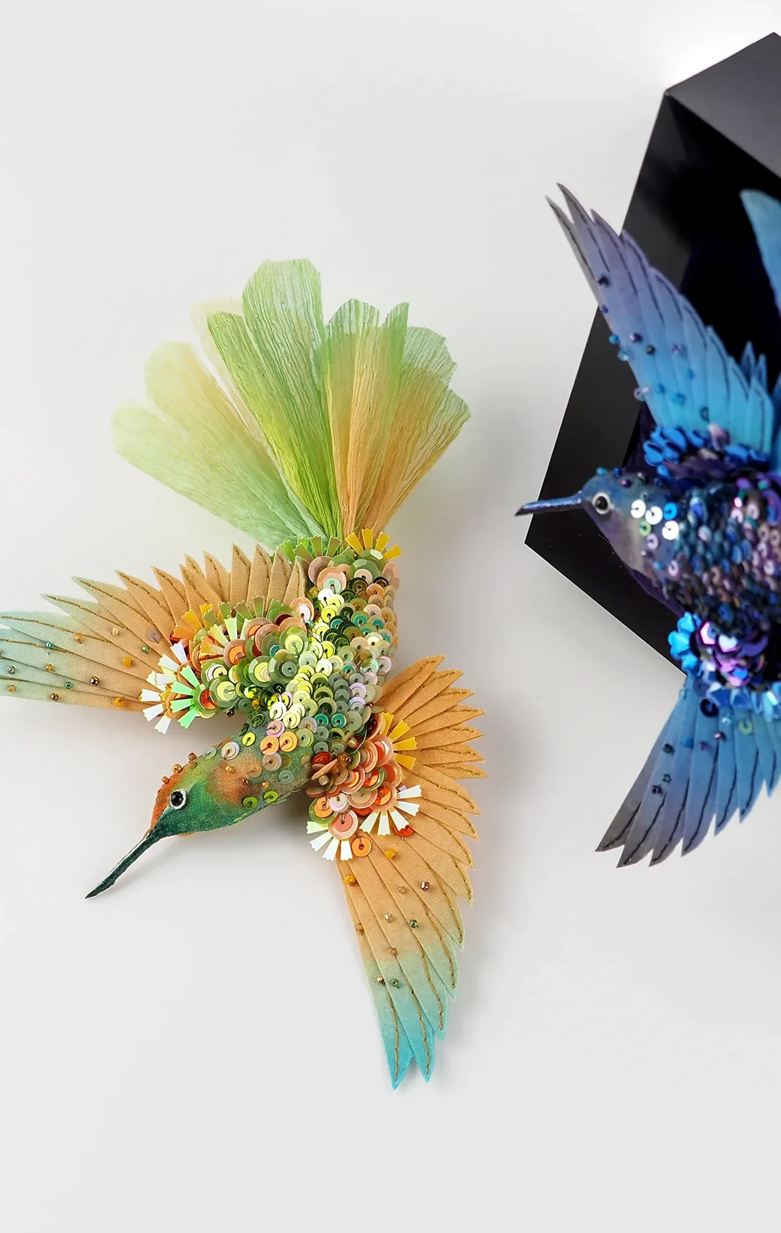 Two handcrafted hummingbird brooches in vibrant green and blue tones, adorned with sequins and beads. One bird is displayed on a velvet frame, the other on a white surface