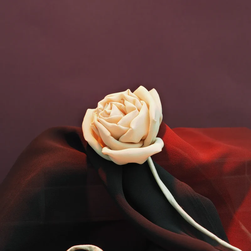 Close-up of a handcrafted silk rose in a soft nude color, artistically displayed on black and red fabric with a rich burgundy background, showcasing intricate folds and lifelike texture.