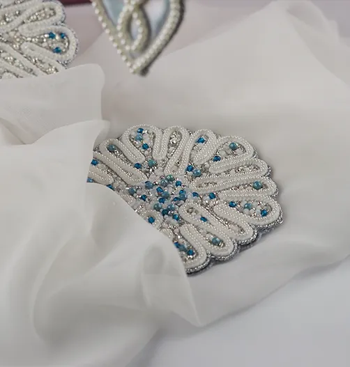 A flat snowflake-shaped embroidered ornament from the Jewels of Winter project, resting on a sheer white fabric. Features white, silver, and blue beads for a luxurious winter theme