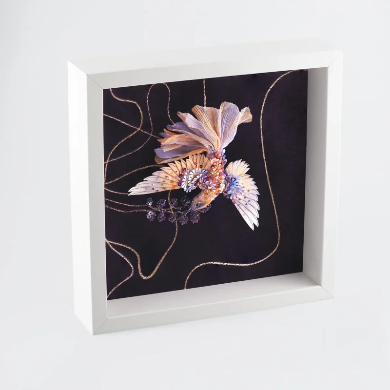 Side view of a luxury framed textile artwork showcasing a meticulously hand-embroidered hummingbird in purple and gold hues. The detailed design features shimmering sequins, delicate stitching, and a deep purple background, framed in a modern white box frame