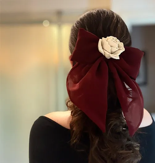 A person with long, wavy brown hair wearing a handcrafted burgundy Chiffon Bow Hair Accessory with an ivory silk rose at the center, styled in a half-up hairstyle. The bow drapes softly, creating a romantic and elegant look.