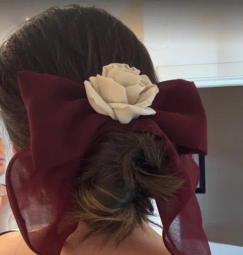 A person with dark hair styled in a low bun, adorned with a burgundy chiffon bow featuring an ivory silk rose at the center