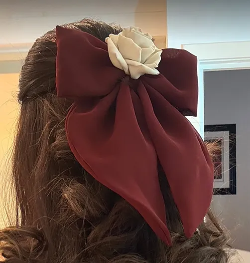 A person with wavy brown hair wearing a handcrafted burgundy or Red Bow Clip for hair with an ivory silk rose at the center, styled in a half-up hairstyle.