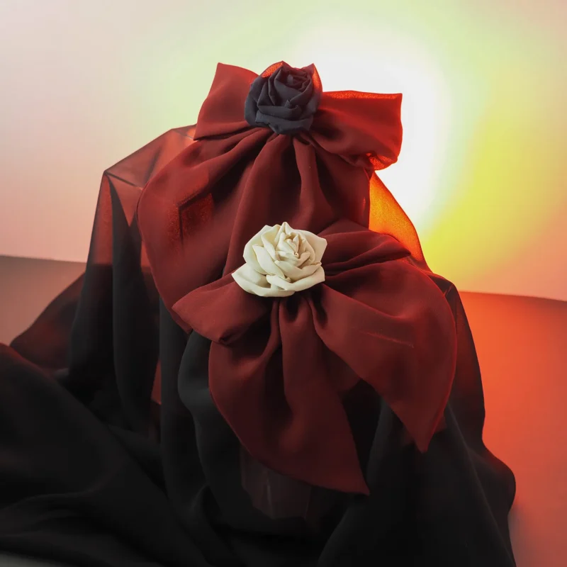 Set of handmade silk bow hair clips in deep red, featuring roses in black and ivory. Elegant and luxurious hair accessories designed for gothic, romantic, or alternative fashion. Styled against sheer black fabric with a dramatic light effect