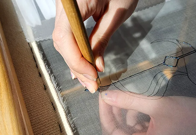 Hand holding a Luneville embroidery hook, stitching black thread along a printed design on sheer fabric stretched in a wooden frame.