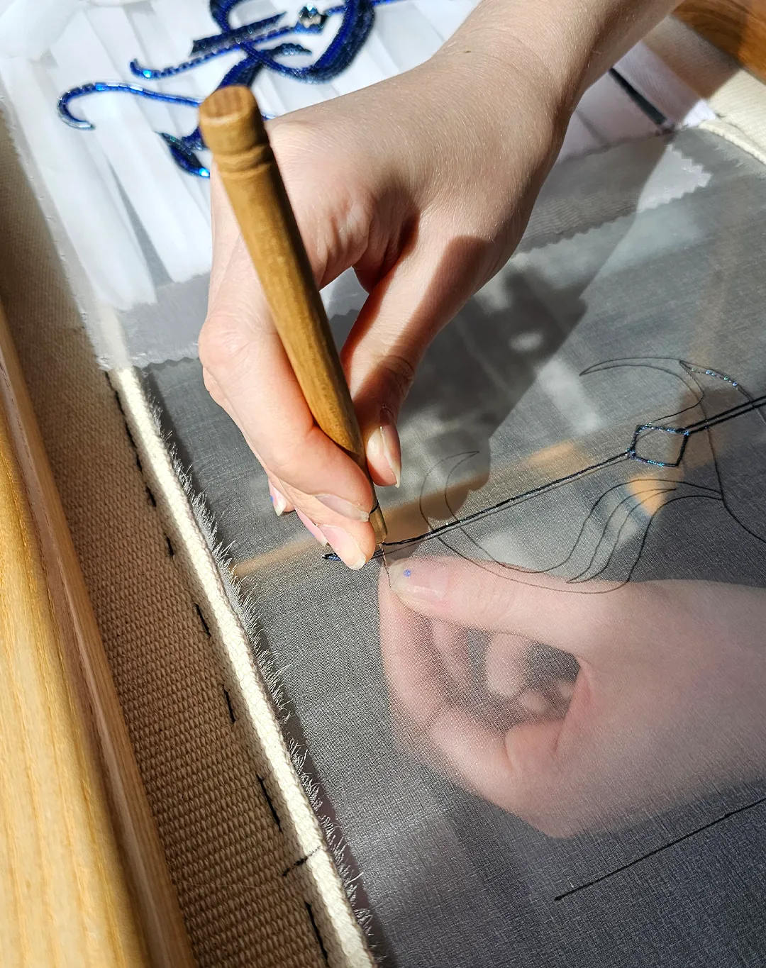 Luneville Embroidery Hook in Use Hand Embroidery on Frame | Ksenia Semirova. Embroidery art Close-up of a hand using a Luneville embroidery hook on sheer fabric stretched in a wooden frame, with blue thread stitching visible