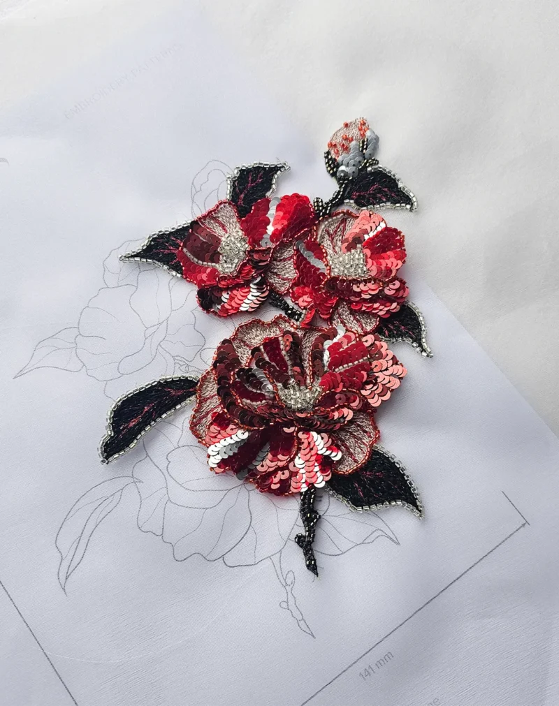 Red and black Luneville embroidery flower with sequins and beads on fabric, placed over a printed embroidery pattern.