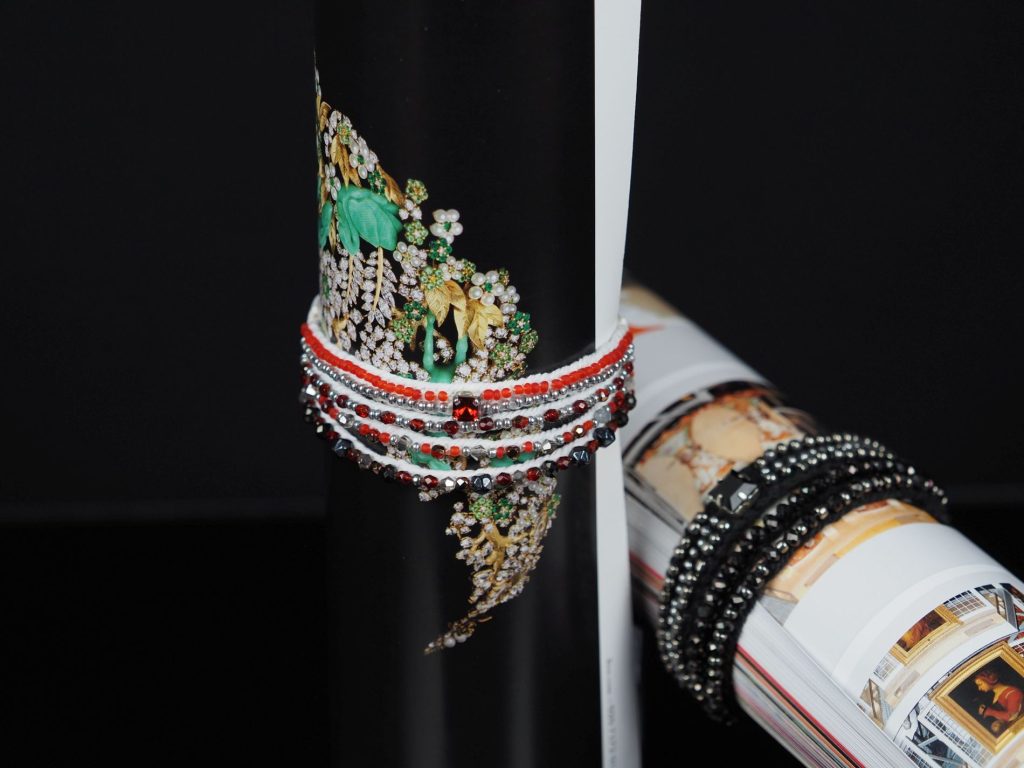 Close-up of a handcrafted beaded bracelet in red, white, and black tones, stitched with pearl setting embroidery and wrapped around a magazine with floral imagery.