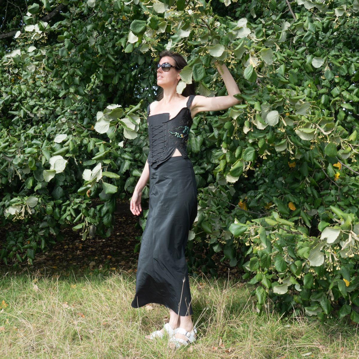 A woman wearing a black embroidered corset with front lacing and side “No” motif, photographed outdoors with hand raised into tree canopy – by Ksenia Semirova