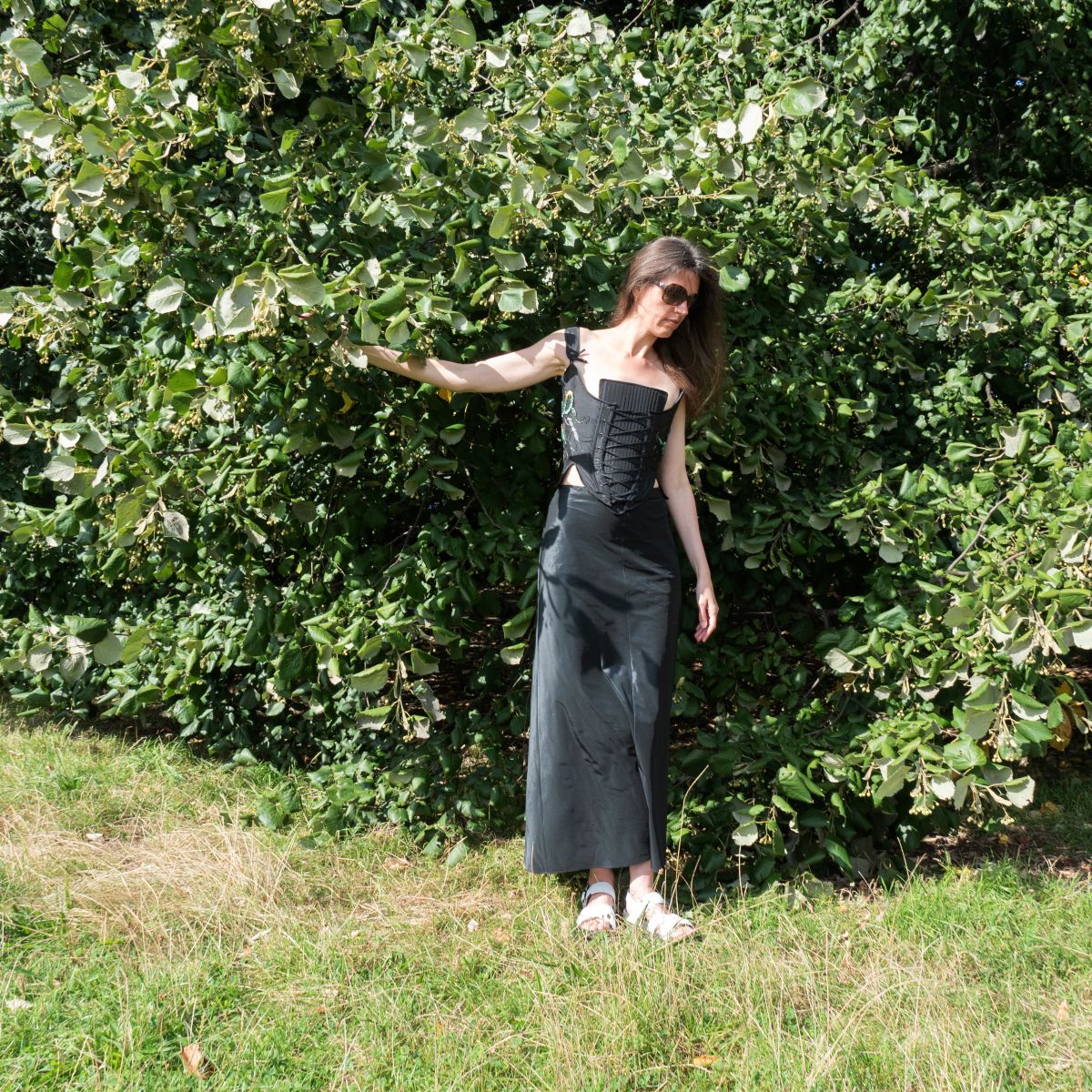 Full-body photo of a woman wearing a black embroidered corset and long skirt, standing in front of green foliage, from The Power of No by Ksenia Semirova