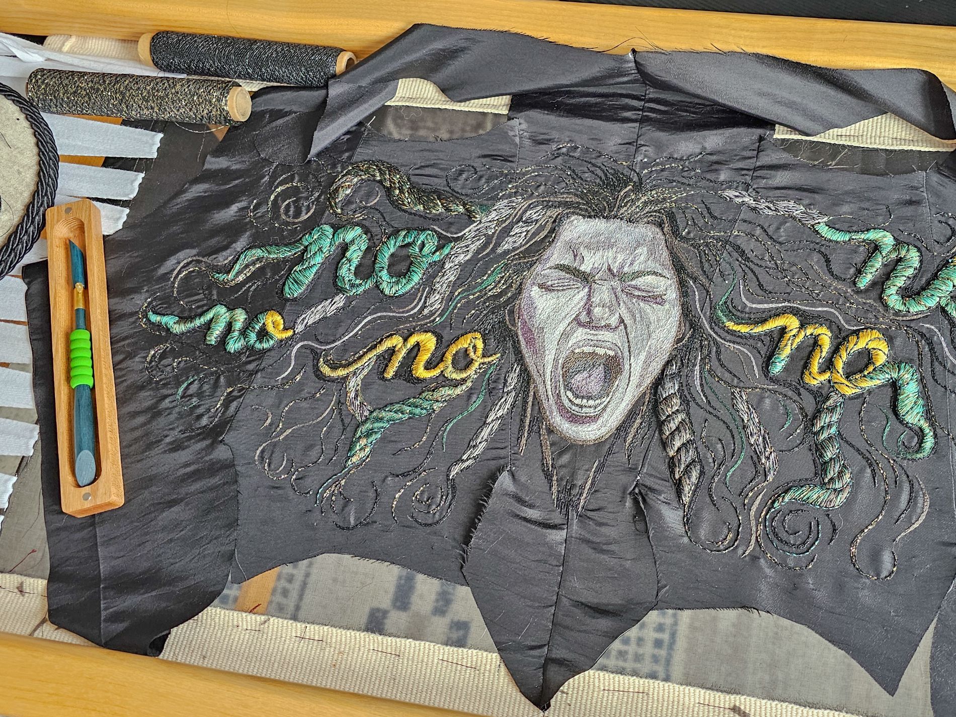 Flat layout showing embroidered screaming face and stitched “No” words in progress, tambour hook and threads visible, from The Power of No corset by Ksenia Semirova
