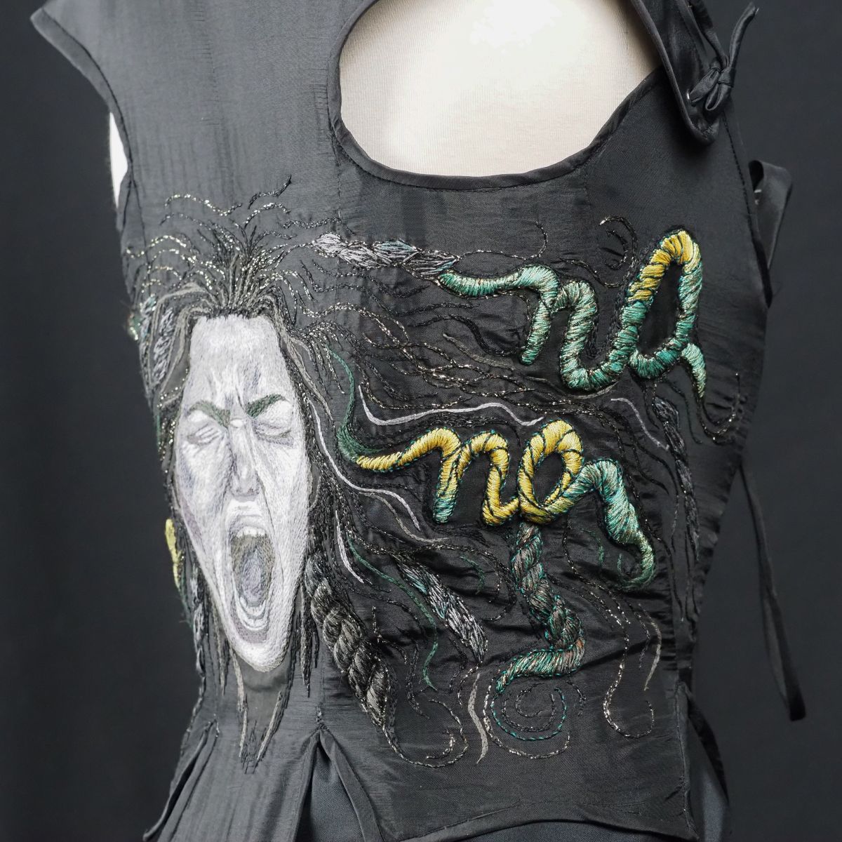Side view of black embroidered corset showing Medusa’s face and textured rope-like hair spelling the word “NO” in green and yellow threads.