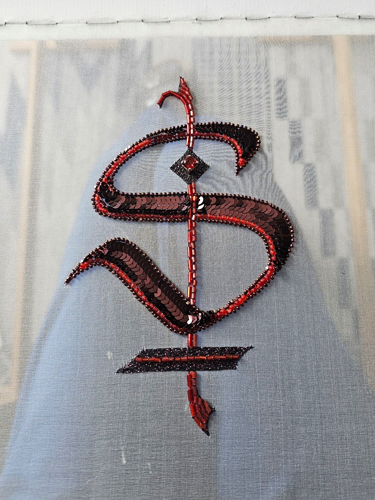 Finished gothic-style monogram embroidered with Luneville and tambour techniques using red and black beads and sequins on sheer fabric, shown stretched on an embroidery frame.