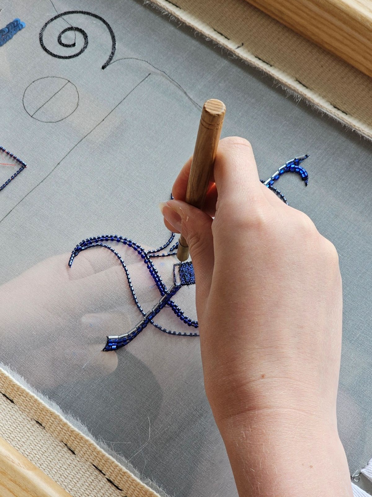 Close-up of Luneville embroidery showing a gothic-style monogram stitched with blue beads on sheer fabric, worked on a wooden embroidery frame using a tambour hook.