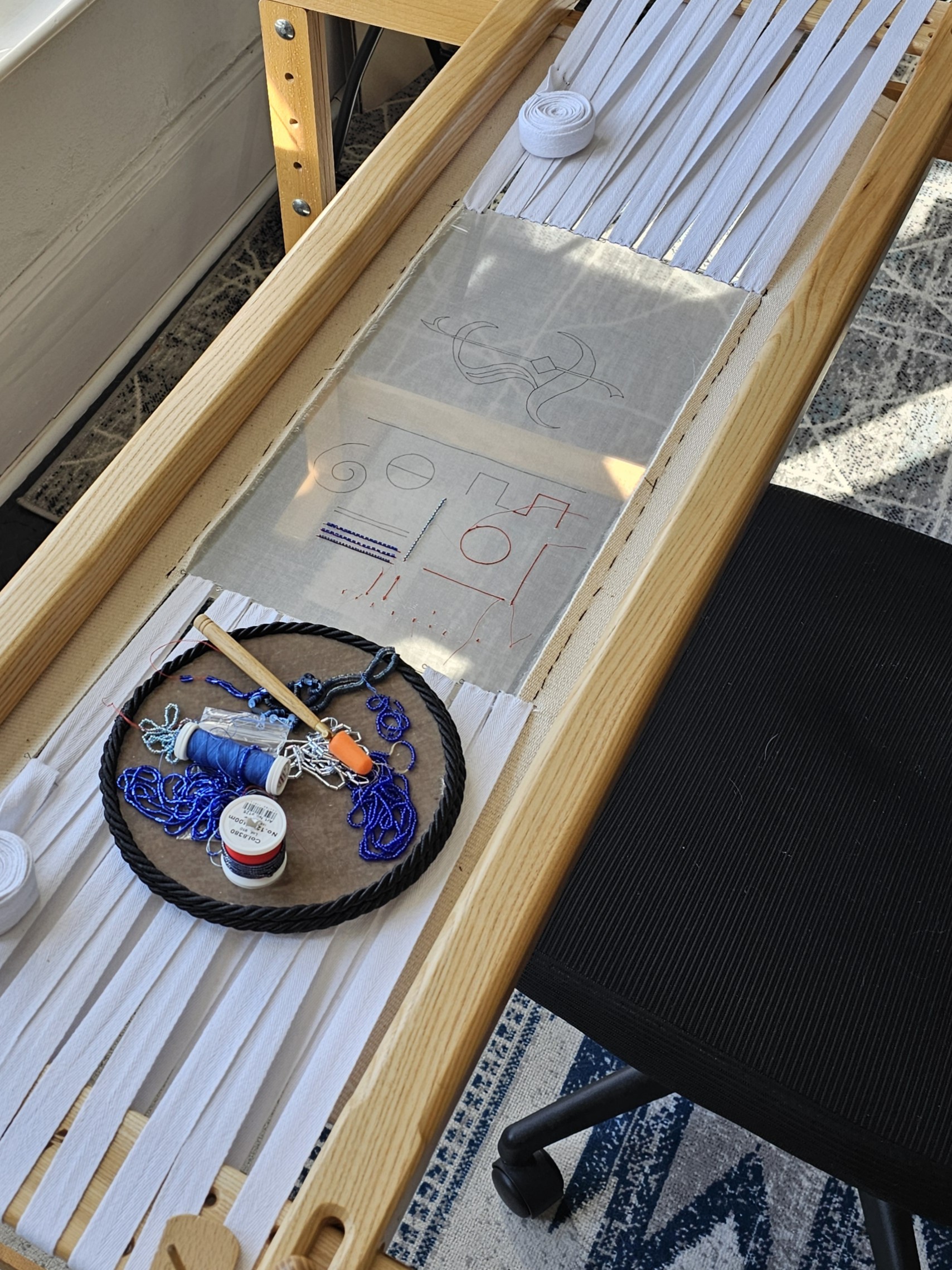 Luneville embroidery setup on a wooden frame showing sheer fabric with drawn practice shapes and a gothic monogram layout, tambour hook, threads, beads, and tools prepared for stitching.