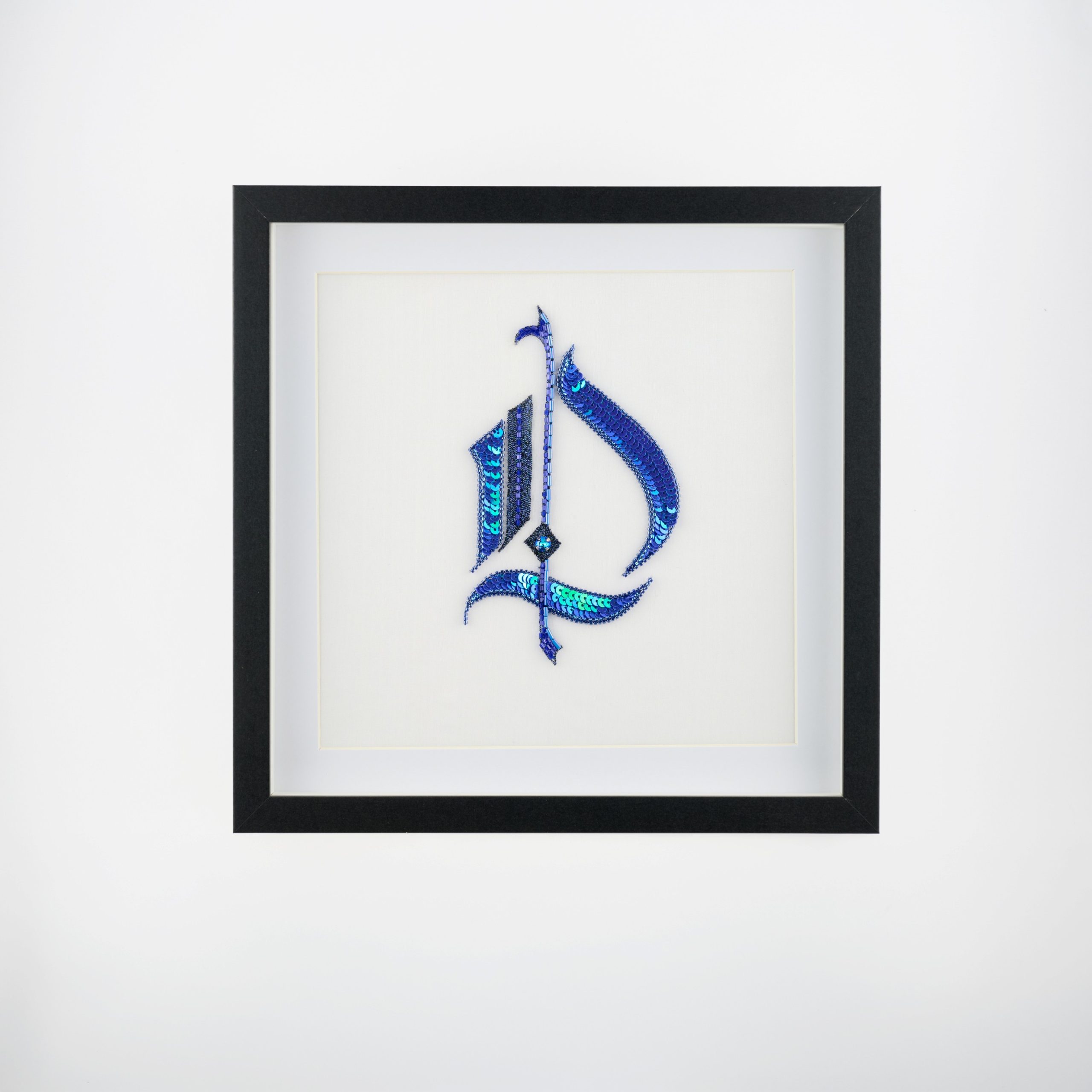 gothic-monogram-wall-art-letter-d-framed-textile-25x25.jpg Gothic monogram wall art featuring the letter D in blue sequins, presented as a framed textile piece in a 25 × 25 cm frame with glass