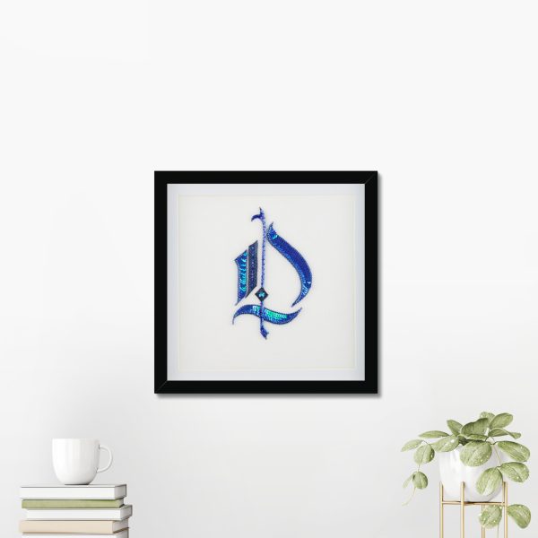 Gothic monogram wall art with the letter D in blue sequins, displayed in a 25 × 25 cm black frame on a wall above a table with books and a plant
