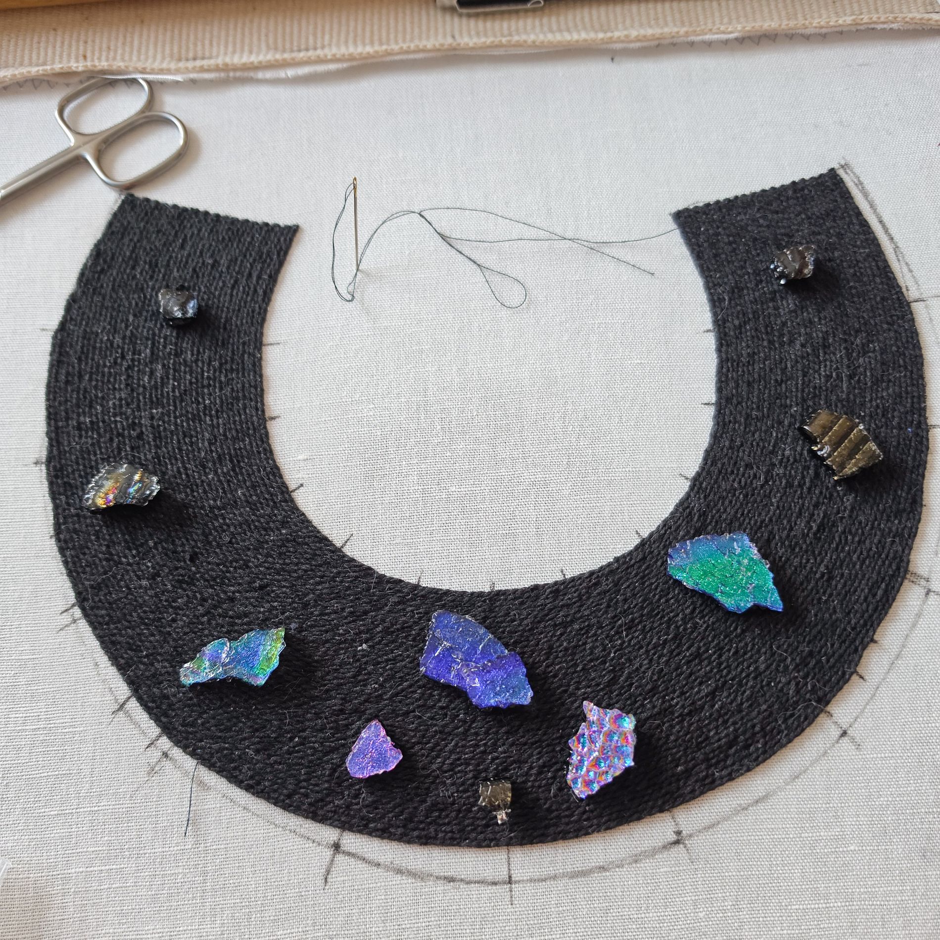Black collar necklace in progress with colourful handmade glass elements arranged across the embroidered base.