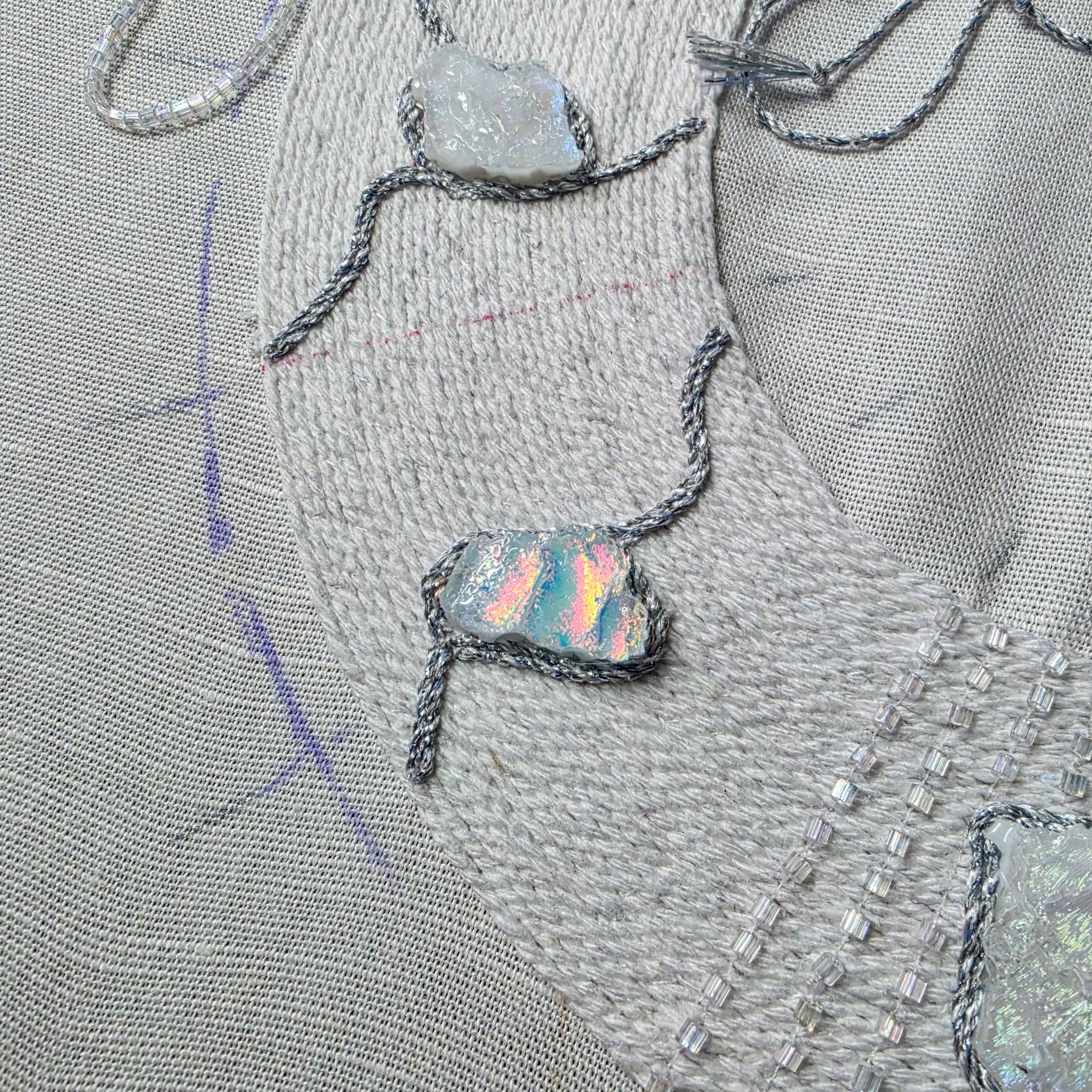 Close-up of an iridescent glass element being secured within the hand embroidery of the white necklace.